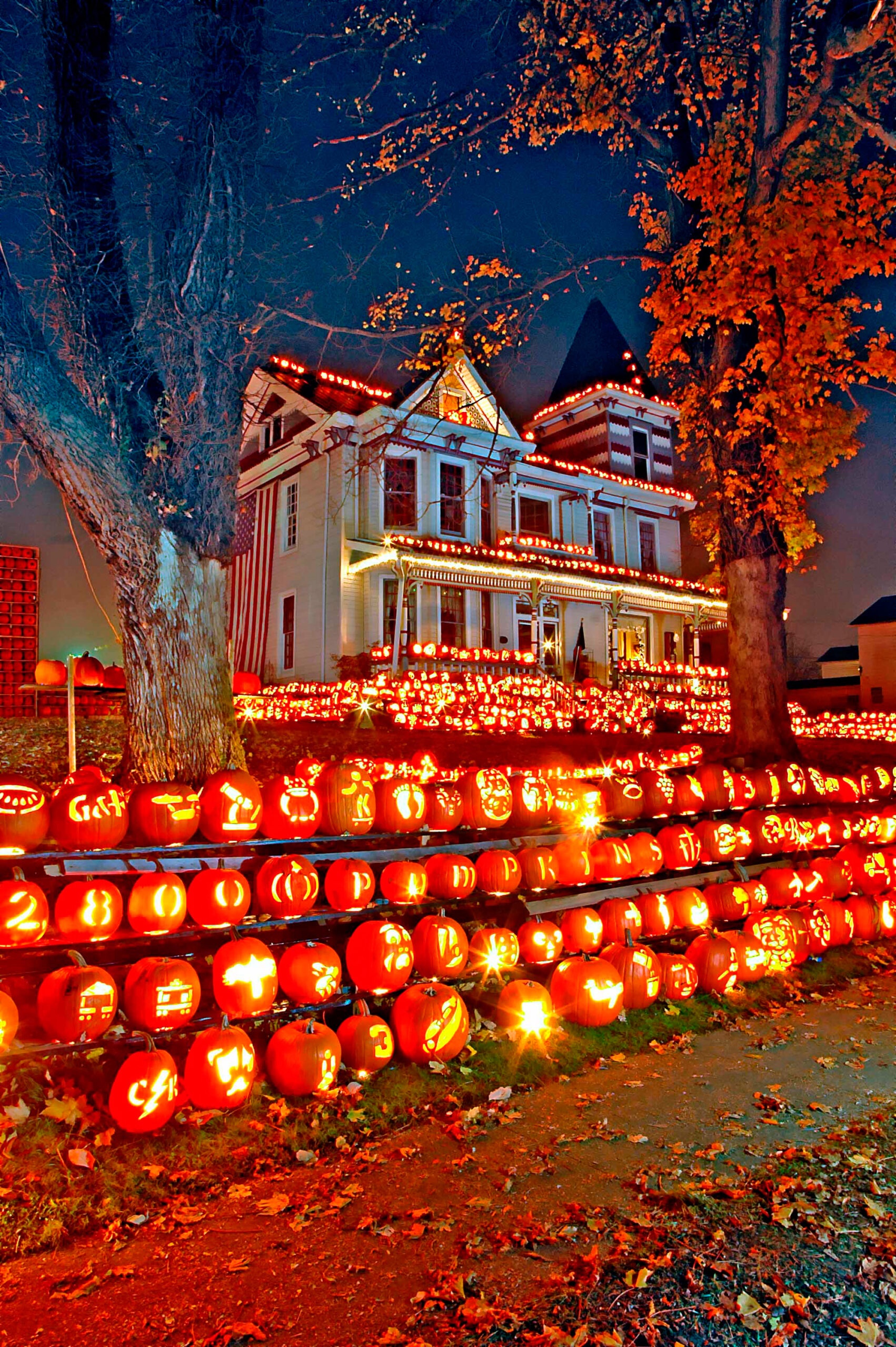 a multi-story house decorated with thousands of glowing carved jack-o'-lanterns on tiered display racks in the front yard at night, flanked by autumn trees
