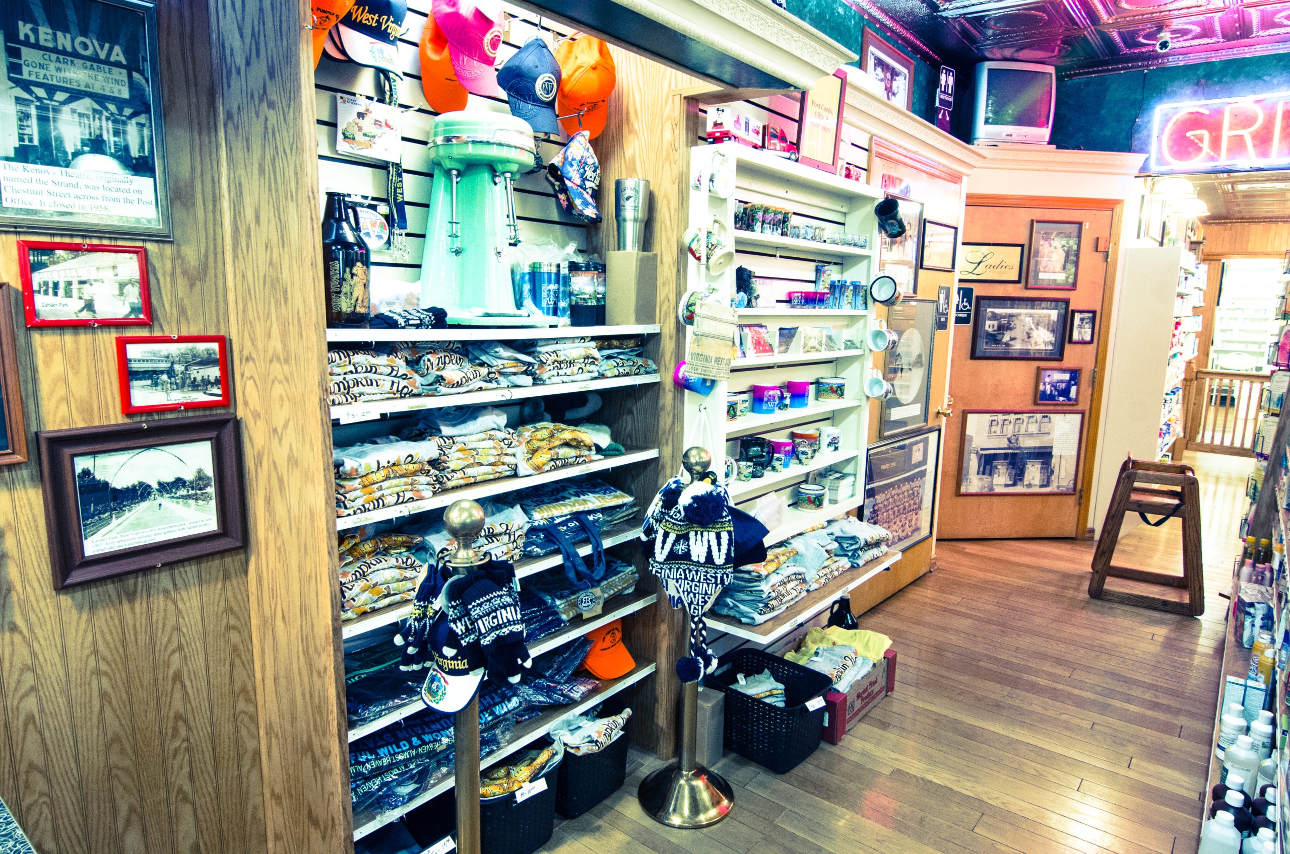 A retro souvenir and gift shop interior with wooden shelves of apparel, hats, a vintage mint-green milkshake mixer, mugs, and framed historical photos under warm lighting
